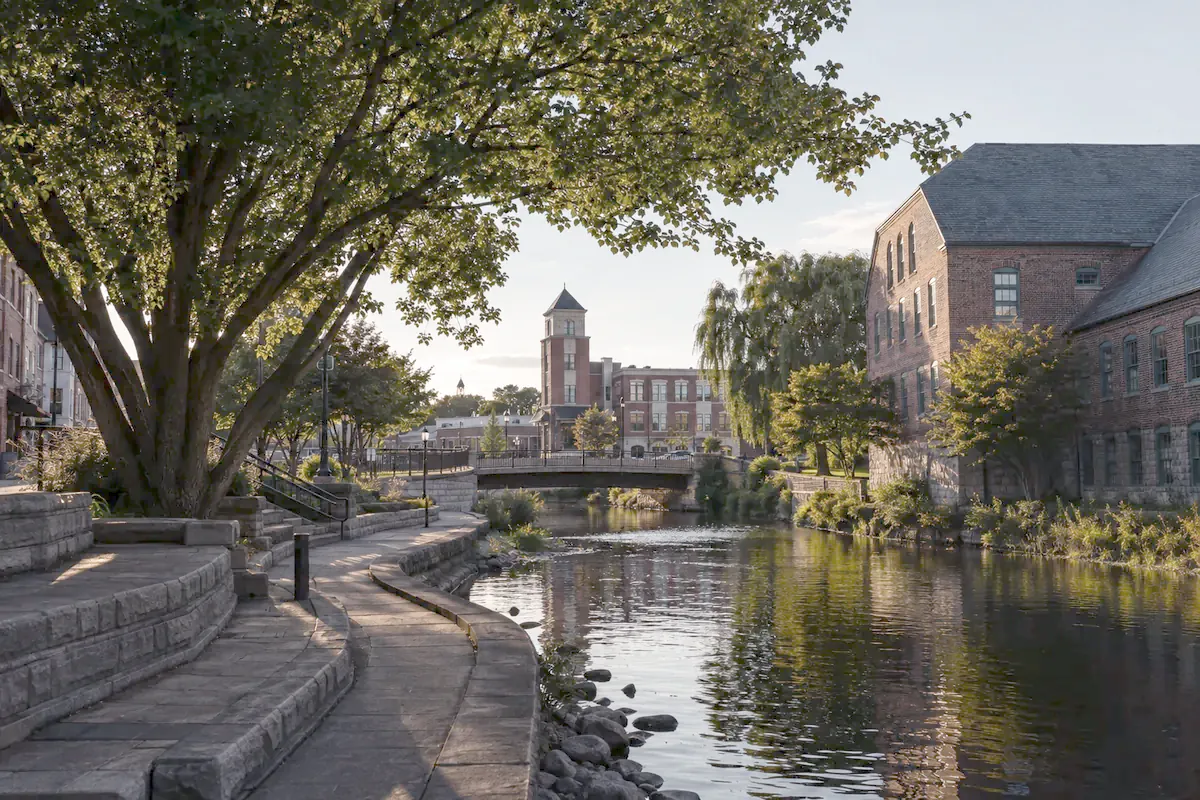 Illustration of West Bend: a curving riverside walkway below trees and brick buildings.